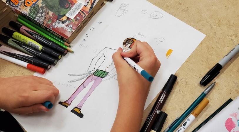 teen hand drawing a character on white piece of paper. markers and books on table.